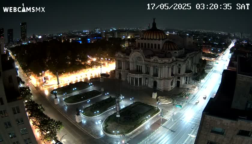 An aerial nighttime view of a large, ornate building illuminated, situated in a city square with a roadway and surrounding parkland under partly cloudy skies.