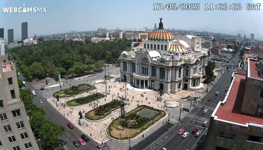 A large, ornate palace-like building sits in a city square with surrounding green landscaping and roads filled with cars, under a clear, sunny sky.