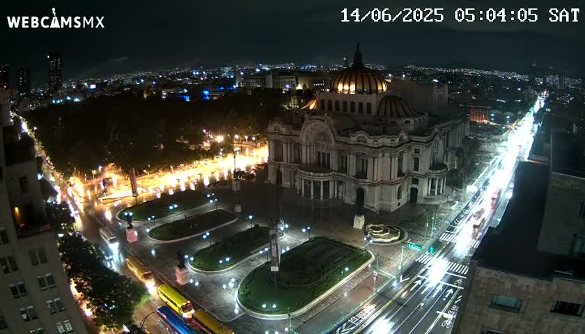 A nighttime aerial view shows a large, ornate building illuminated, surrounded by a plaza with landscaping and roadways filled with traffic under a clear night sky.