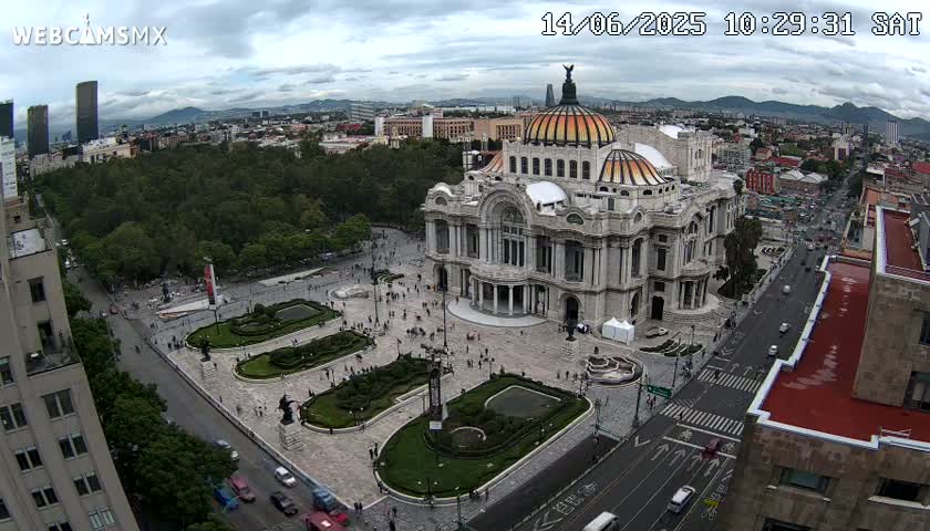 An aerial view shows the Palacio de Bellas Artes in Mexico City on an overcast day, surrounded by a plaza and city streets with sparse traffic.