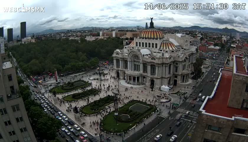 An aerial view shows the Palacio de Bellas Artes in Mexico City on an overcast day, surrounded by a plaza and city streets with sparse traffic.