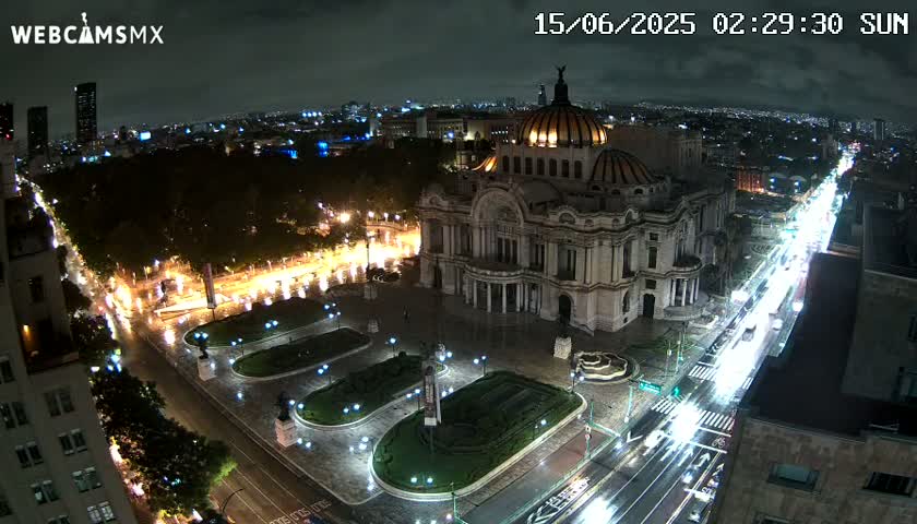 A nighttime aerial view of a large, ornate building surrounded by a plaza with landscaped areas and a busy street with car lights.
