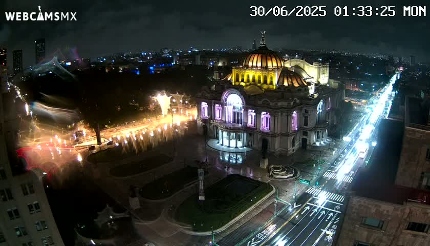 A nighttime aerial view shows a large, illuminated building, possibly a museum, situated in a city square with fountains, and a busy street with traffic flowing past it under a partly cloudy sky.