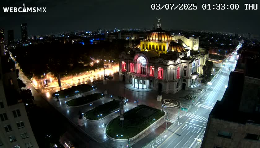 An illuminated palace-like building sits in a plaza at night, with a sparsely trafficked street extending from it into a city skyline under a clear sky.