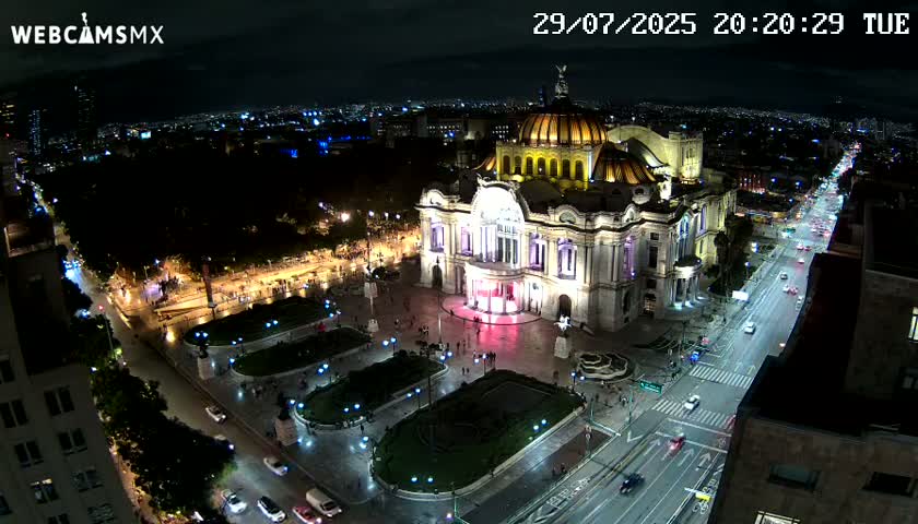 A brightly lit palace-like building stands in a plaza at night, with a city street extending from it, sparsely populated with cars under clear skies.