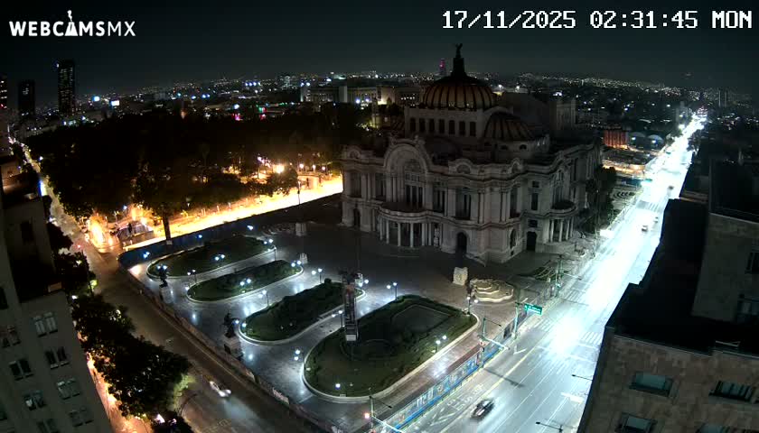 A grand, ornate domed building is brightly illuminated at night, flanked by a busy city street with light trails from vehicles and a well-lit park, all under clear skies.