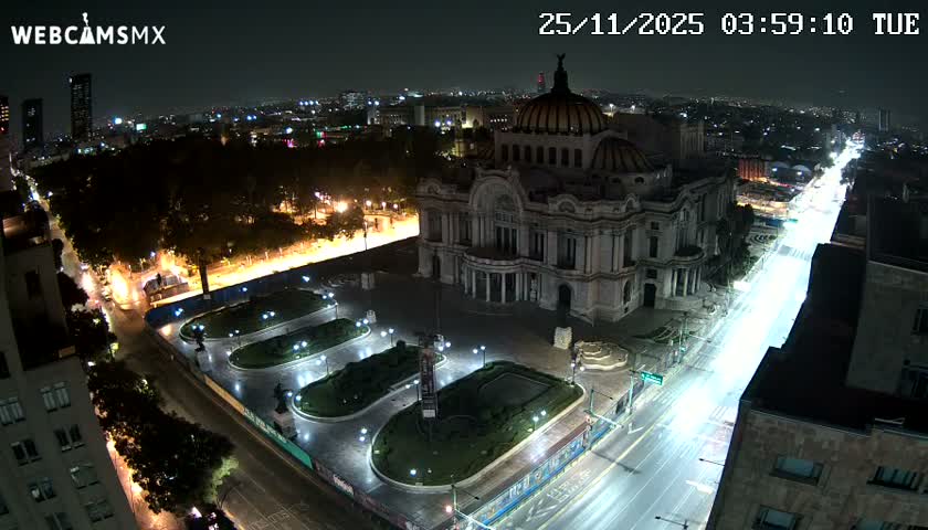 A brightly lit, ornate domed building stands prominently at night in an urban setting, flanked by a park with illuminated pathways and a busy street featuring long streaks of vehicle lights, all under a clear sky.