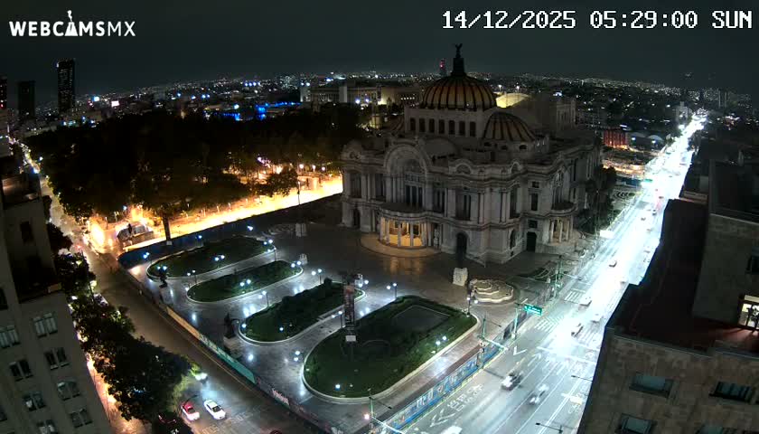 A brightly lit, ornate domed building stands prominently at night in an urban setting, flanked by a park with illuminated pathways and a busy street featuring long streaks of vehicle lights, all under a clear sky.