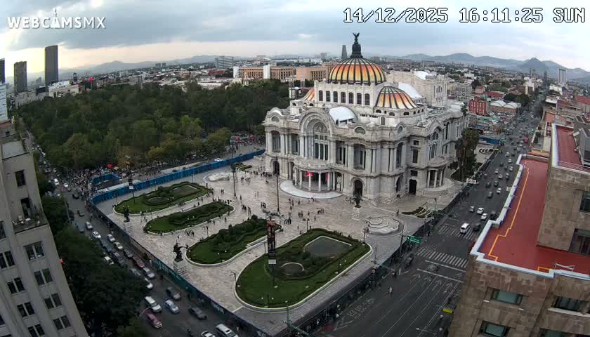 A brightly lit, ornate domed building stands prominently at night in an urban setting, flanked by a park with illuminated pathways and a busy street featuring long streaks of vehicle lights, all under a clear sky.