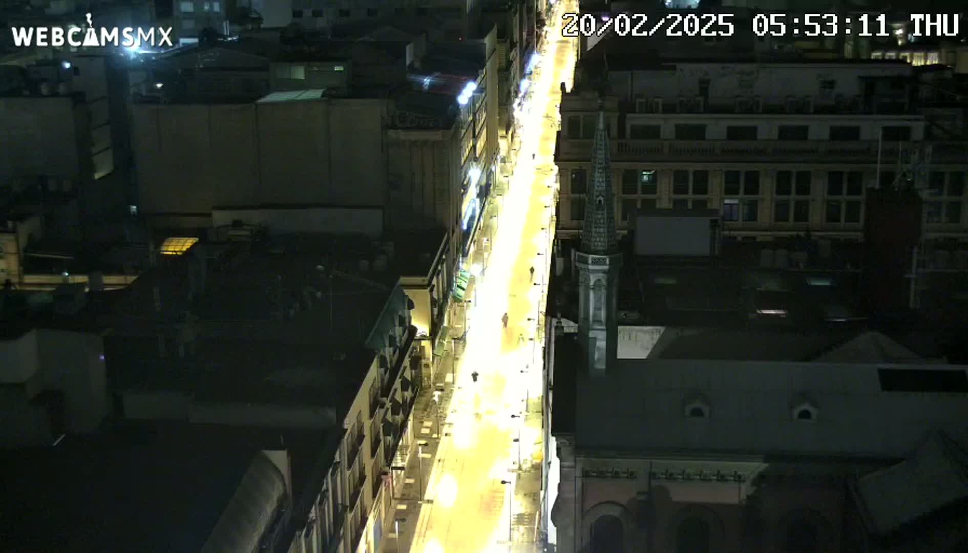 A brightly lit city street at night, viewed from above, shows buildings and few pedestrians.