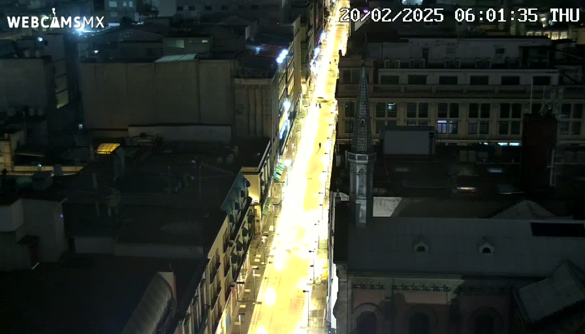 A brightly lit city street at night, seen from an elevated viewpoint, shows buildings on either side with sparse pedestrian traffic.