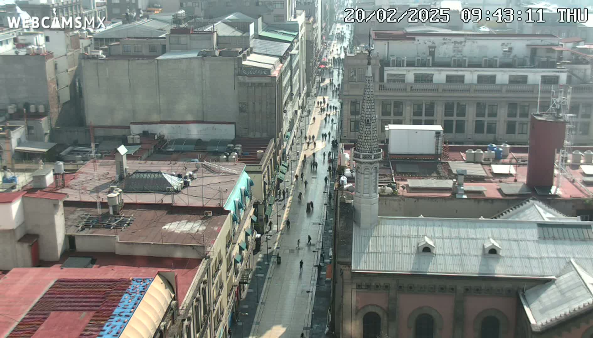 A city street scene on an overcast day shows a long street with pedestrians walking and buildings lining both sides.