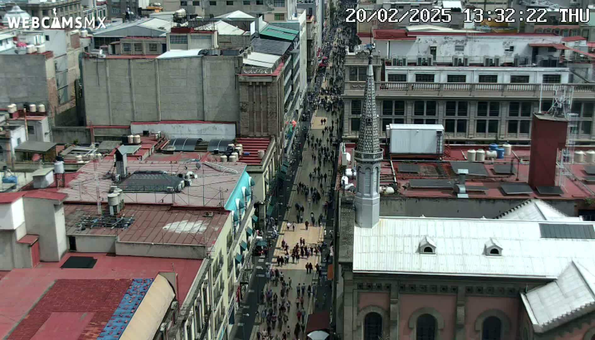 A crowded city street is viewed from above, showing pedestrians walking between buildings on a mostly sunny day.