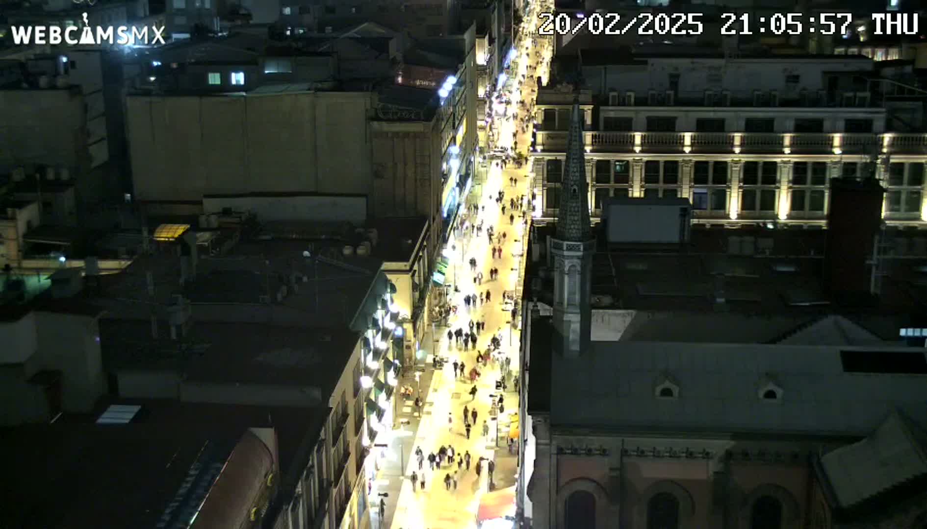 A brightly lit, pedestrian-filled street at night is viewed from above, surrounded by dark buildings.