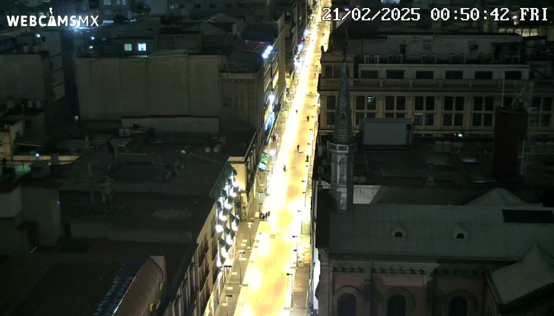A brightly lit, mostly empty street at night is viewed from above, with buildings lining both sides.