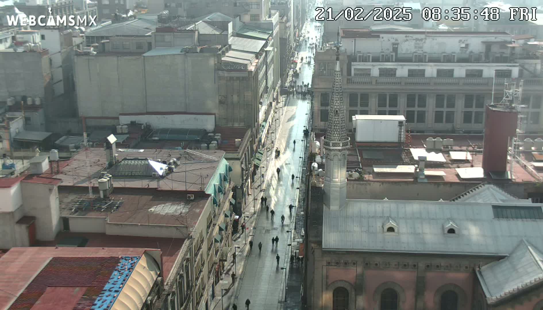 A high-angle, long shot of a city street on a partly sunny day, showing people walking on a wet, paved street lined with buildings.
