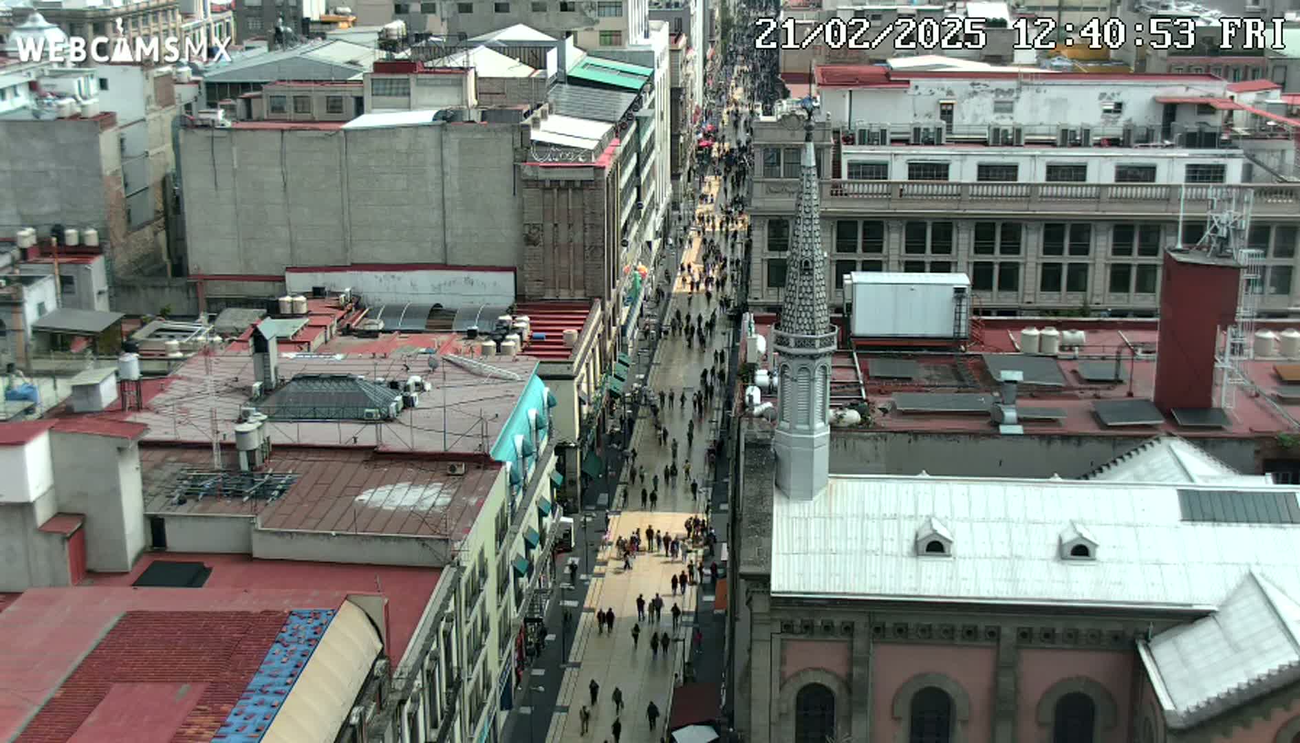 A high-angle view of a city street crowded with pedestrians, flanked by buildings with red tile roofs under an overcast sky.