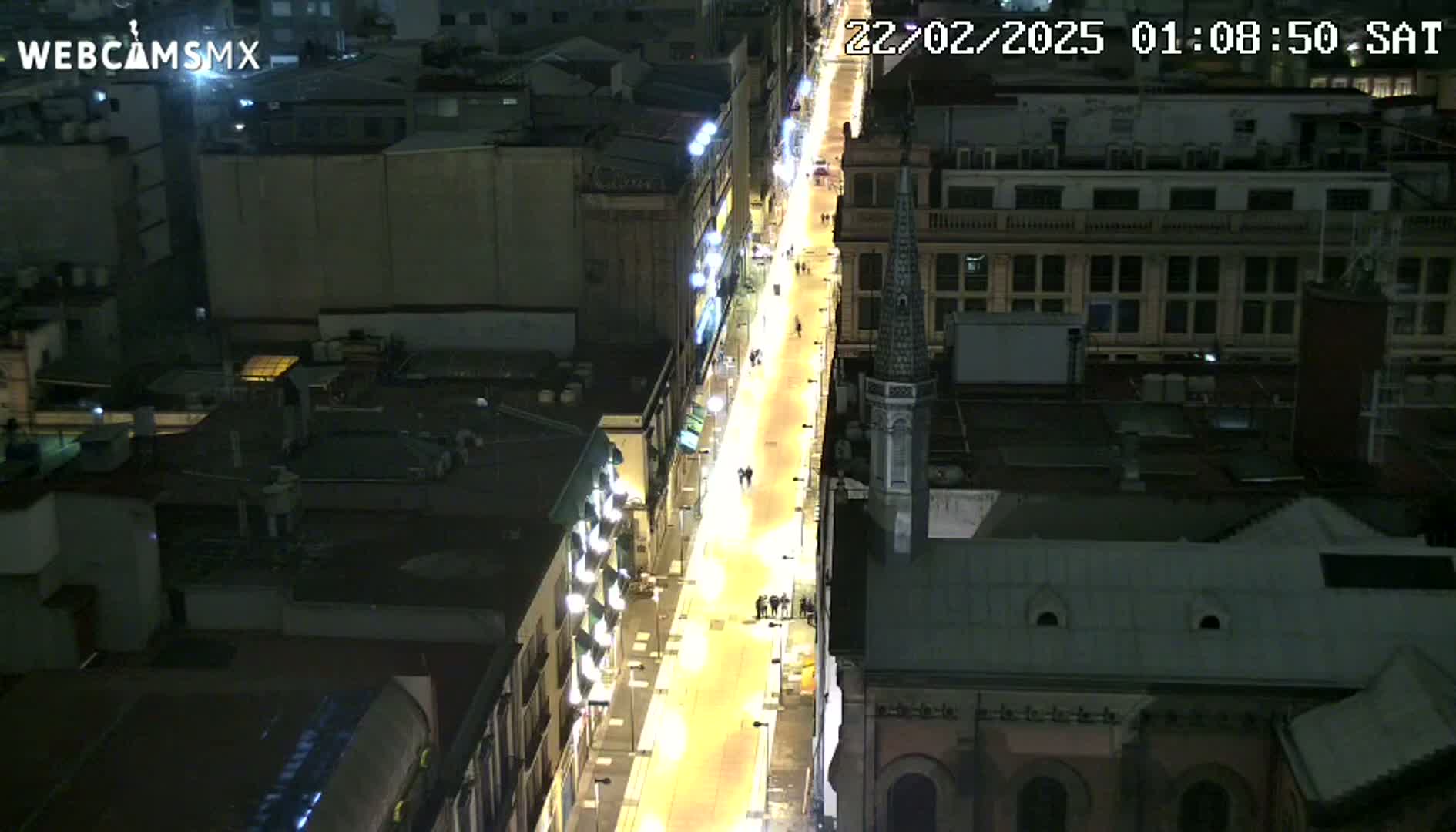 A brightly lit city street at night, viewed from above, shows several people walking along a pedestrianized area between buildings.