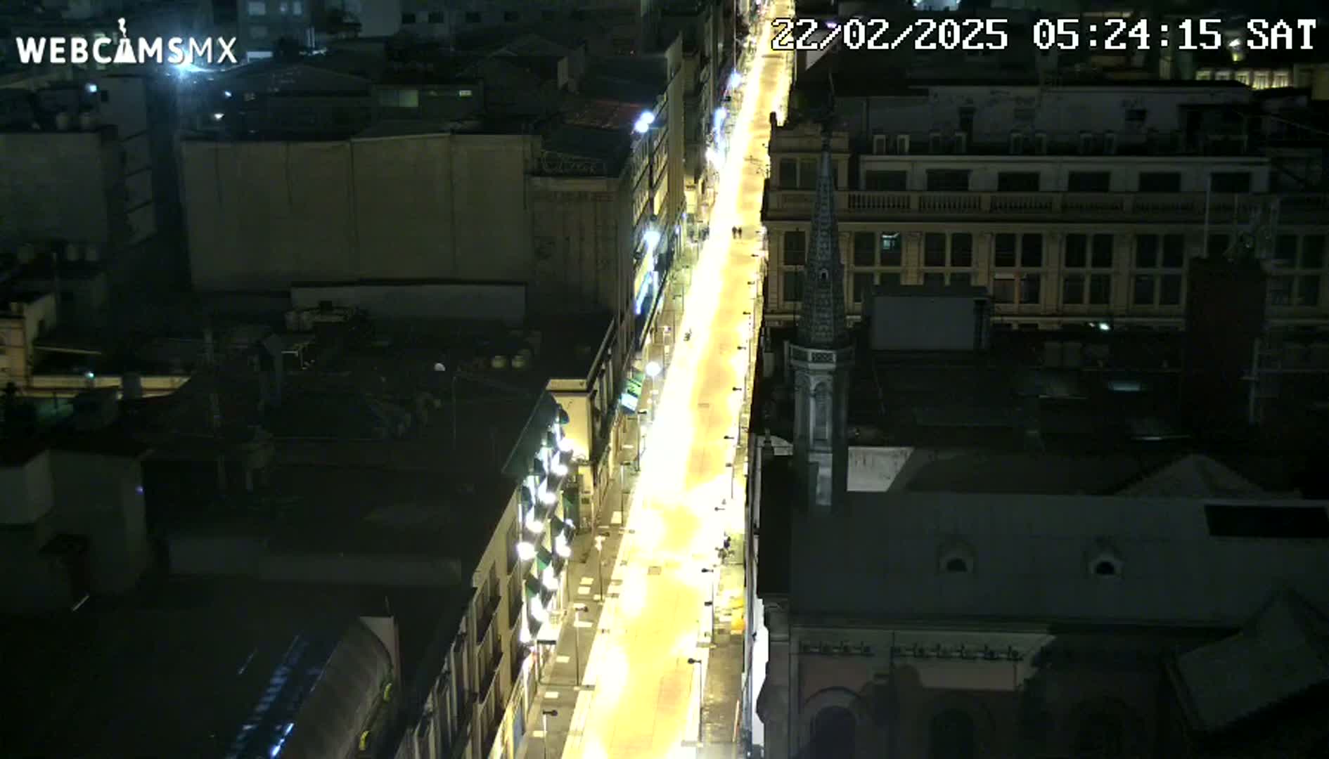 A brightly lit street at night is seen from an aerial perspective, with buildings on either side.