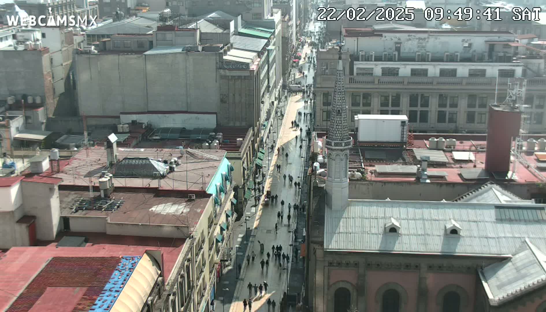 A high-angle view of a city street on a sunny day, showing pedestrians walking along a wide avenue lined with buildings, some of which are multi-story and others appear to be churches or older buildings.