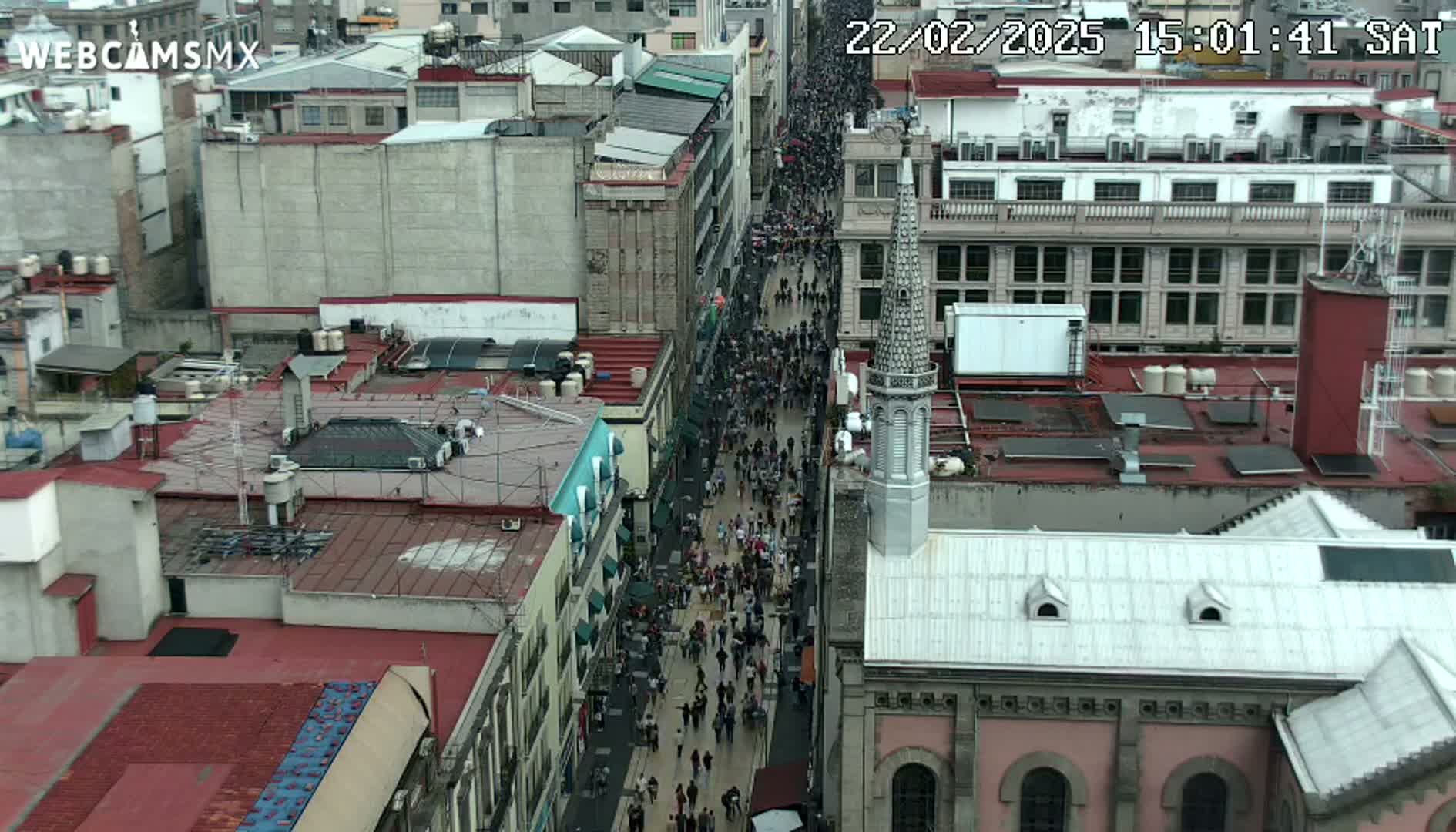 A crowded street scene is viewed from above, showing a city block filled with buildings and many pedestrians walking below on a cloudy day.
