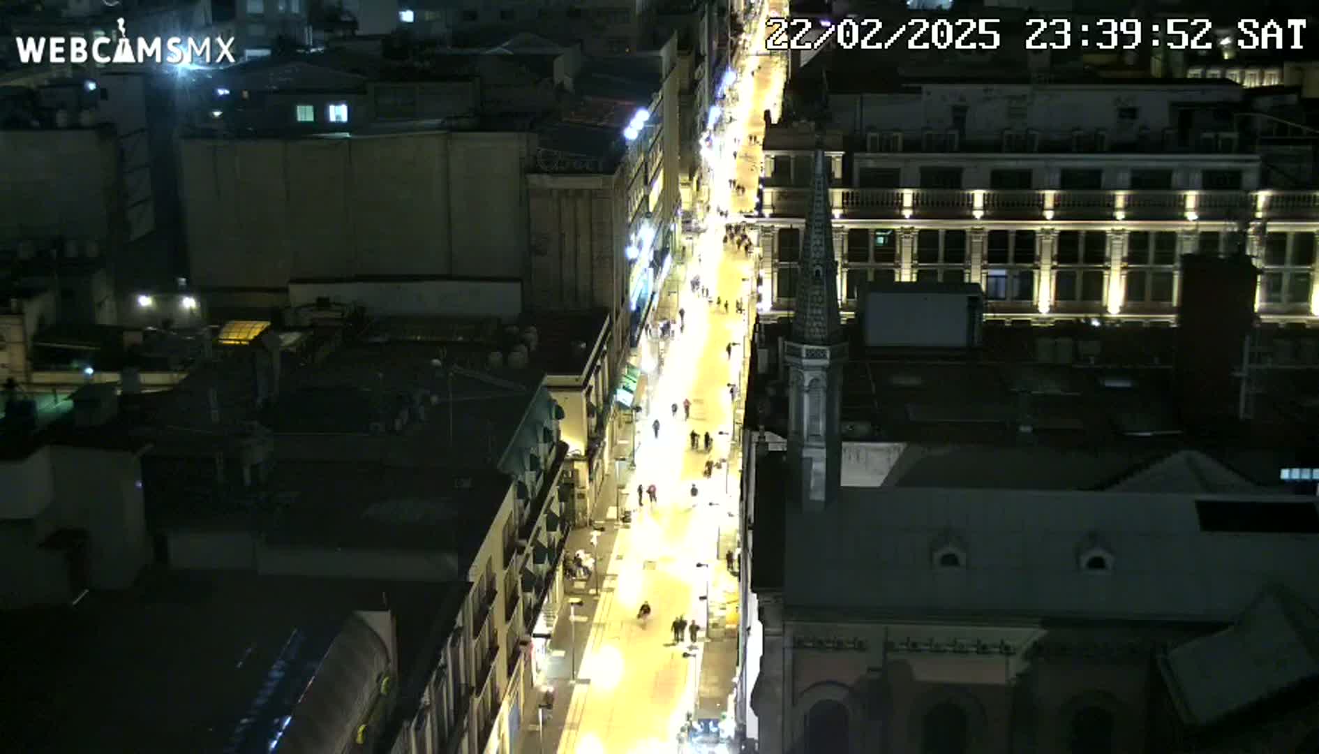 A brightly lit city street at night, filled with pedestrians, is viewed from above.