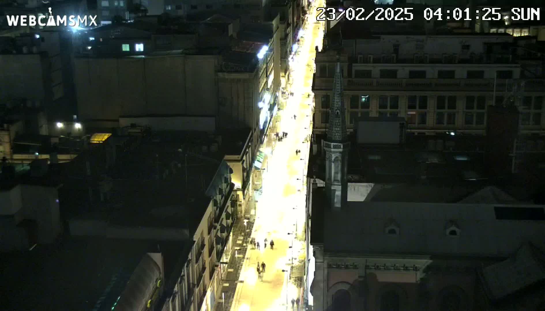 A nighttime, high-angle view of a brightly lit city street, showing buildings on either side and pedestrians walking along the thoroughfare.