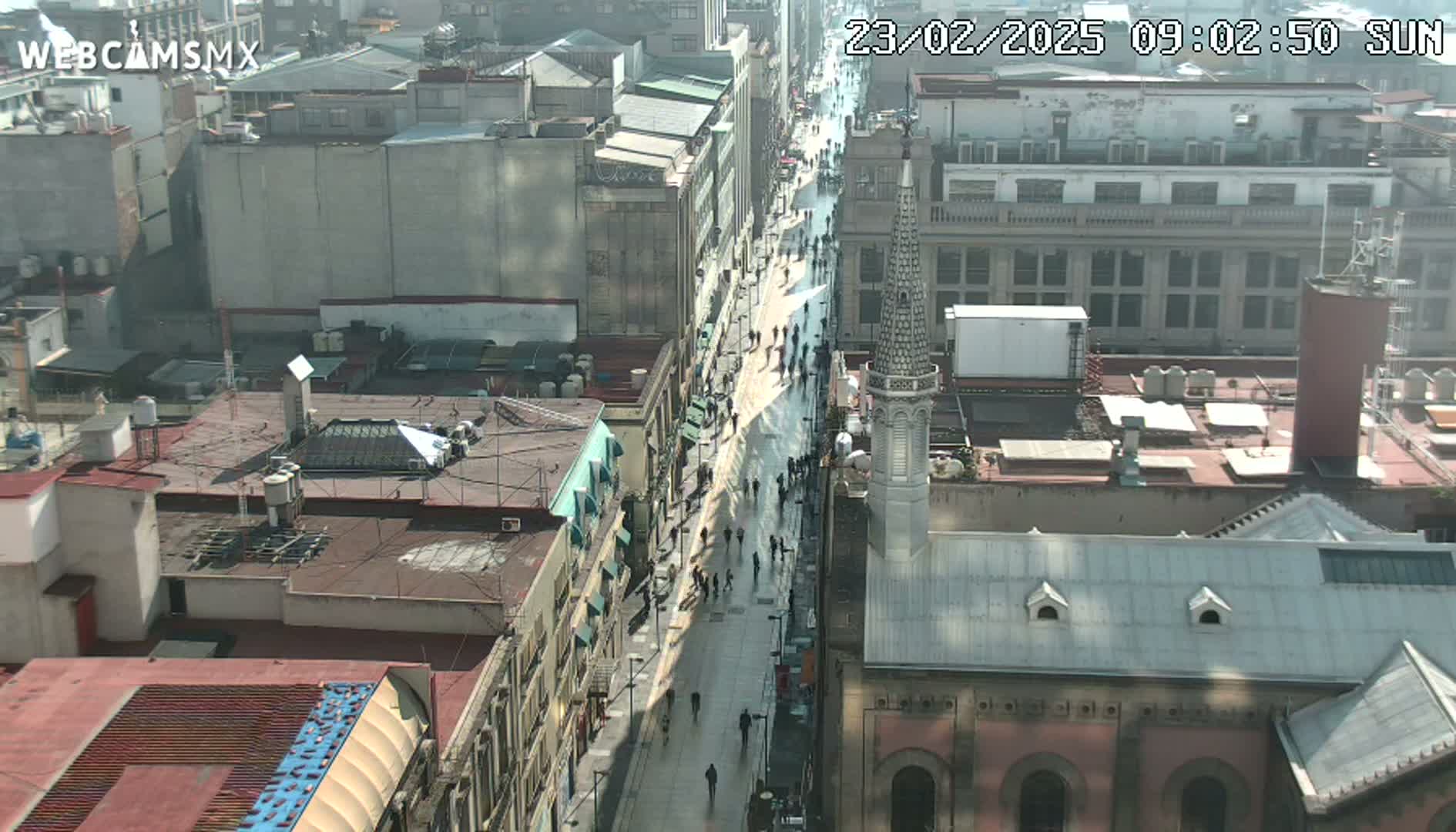 A sunny, high-angle view of a city street with pedestrians, flanked by buildings, one of which has a tall, pointed spire.