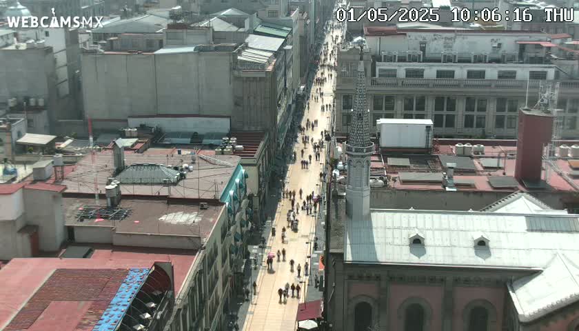 A high-angle, long shot shows a city street crowded with pedestrians on a sunny day, with buildings on either side and a church's roof visible in the foreground.