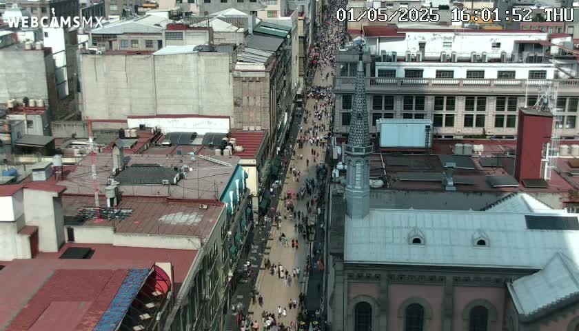 A crowded city street is viewed from above on a sunny day, with buildings lining both sides of the street.