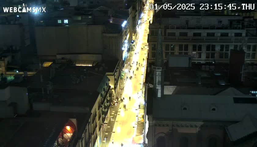 A brightly lit street at night, viewed from above, shows people walking along a pedestrianized area lined with buildings.
