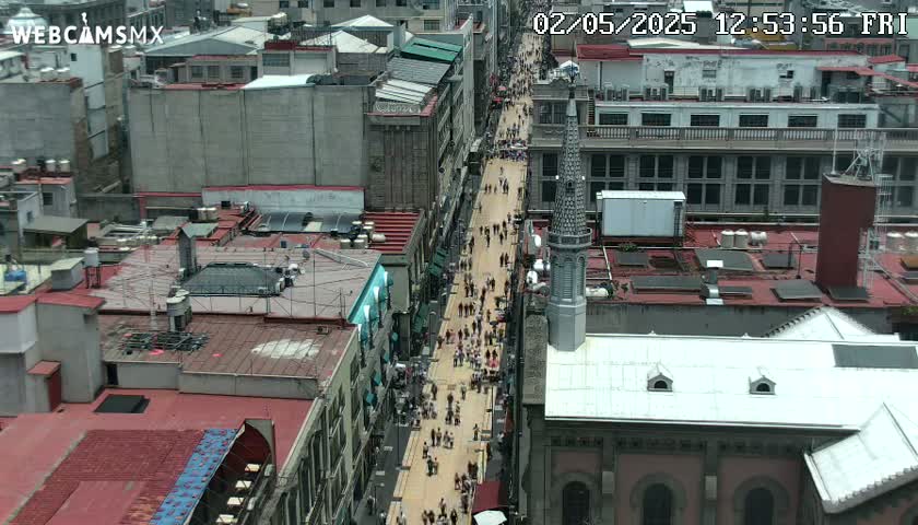 A crowded pedestrian street is seen from above, surrounded by densely packed city buildings under a clear sky.