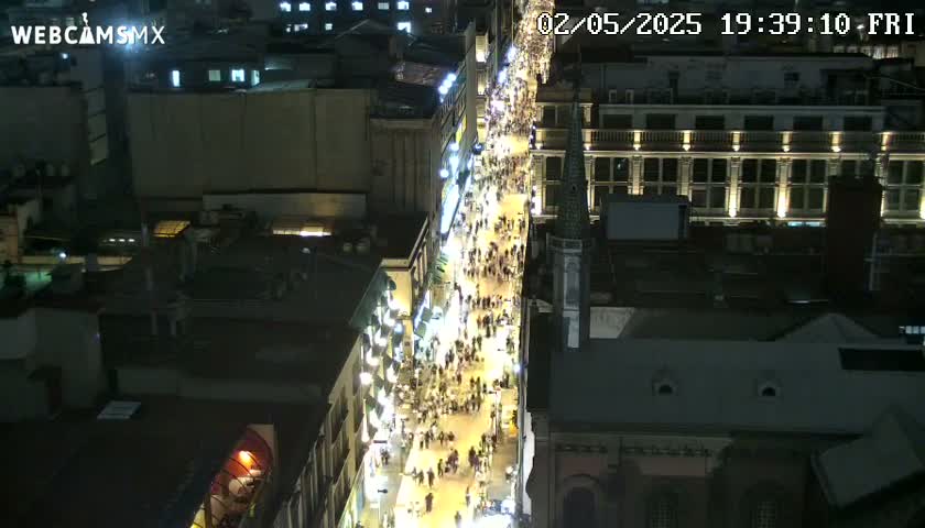 A brightly lit city street at night is crowded with pedestrians, viewed from an elevated perspective.