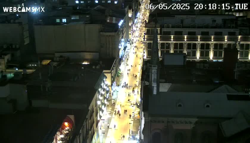 A brightly lit, bustling city street at night is viewed from above, showing many people walking along the sidewalks.