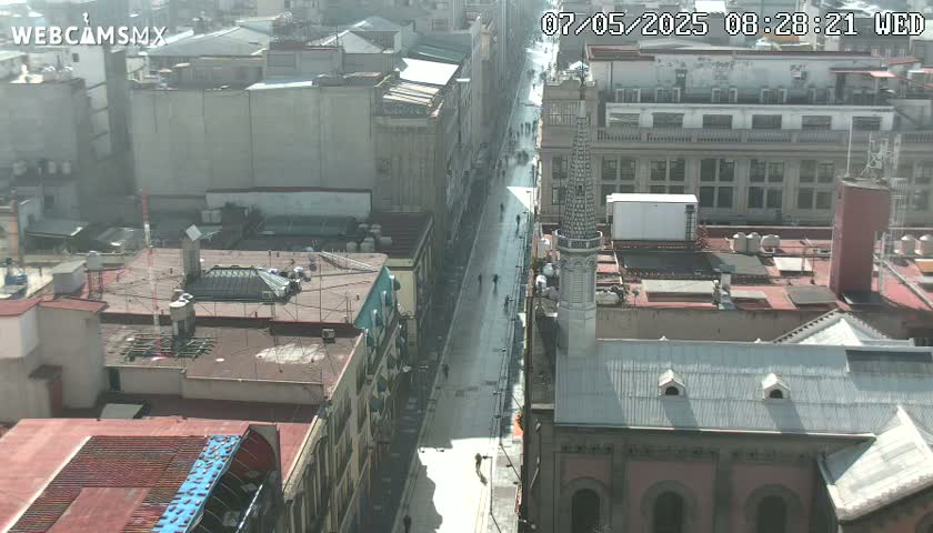 A long city street, mostly empty except for a few pedestrians, is viewed from above on a partly cloudy day.