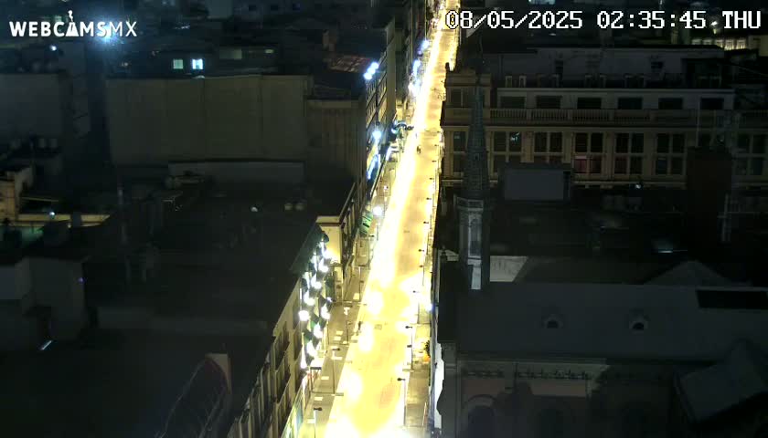 A brightly lit city street at night is viewed from above, showing buildings on either side.