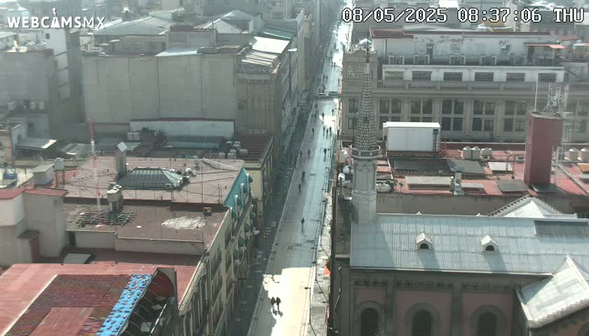 A hazy, sunny day view overlooking a city street with several pedestrians and buildings, including a tall, slender tower.