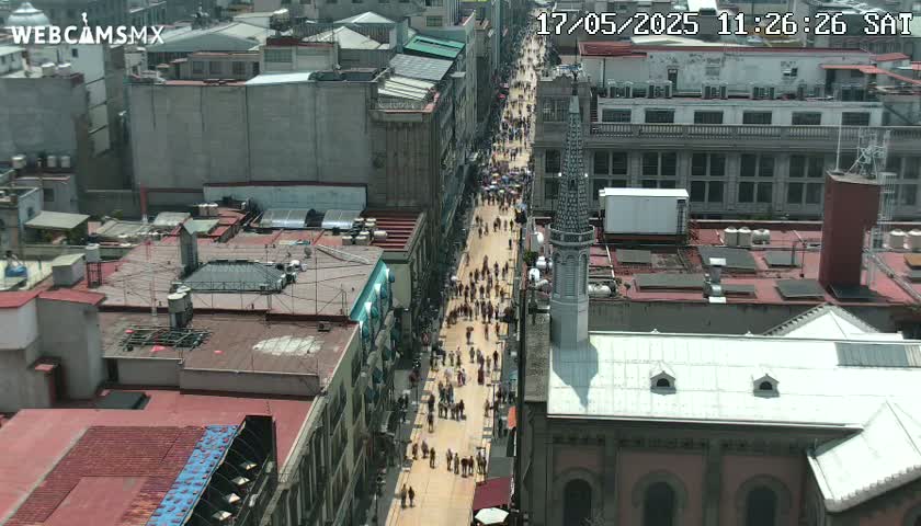A street filled with people is viewed from above on a sunny day, flanked by tall buildings and a church.