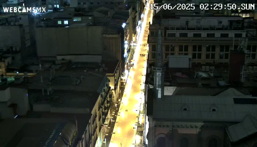 A brightly lit street at night, surrounded by dark city buildings, is viewed from above.