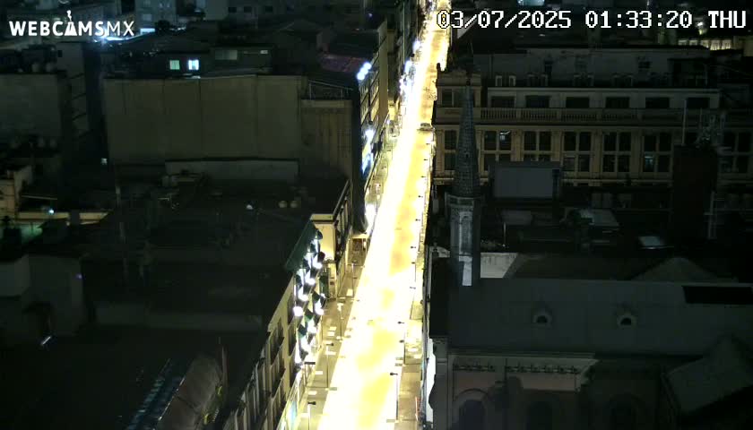 A brightly lit street at night, viewed from above, is flanked by buildings.