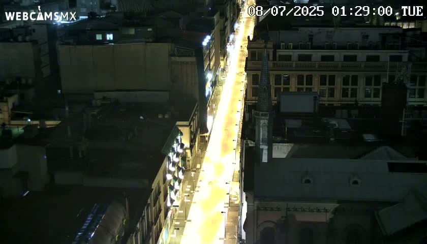 A brightly lit city street at night is viewed from above, showing buildings on either side.