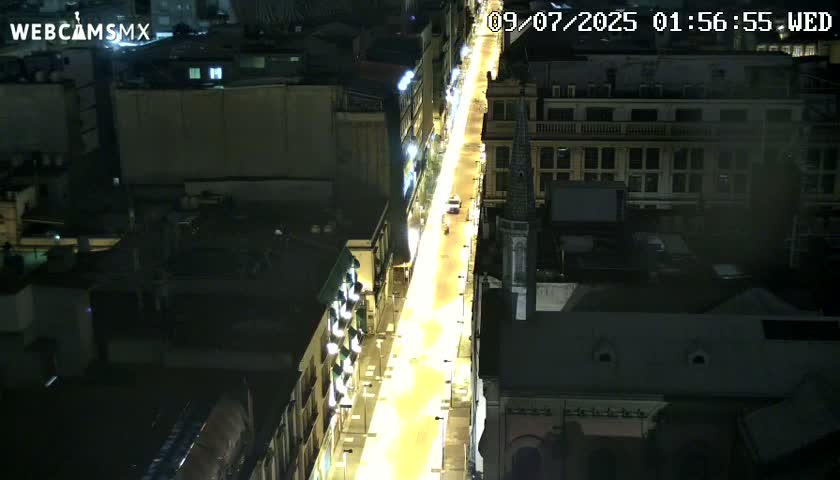 A brightly lit city street at night is seen from an aerial perspective, with buildings flanking both sides of the road.