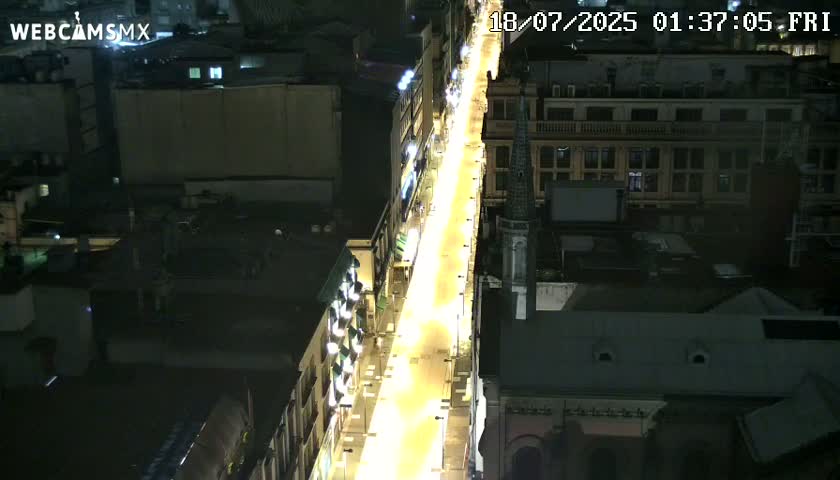 A brightly lit city street at night, viewed from above, showing buildings on either side.
