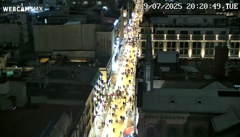 A brightly lit city street at night, viewed from above, showing buildings on either side.