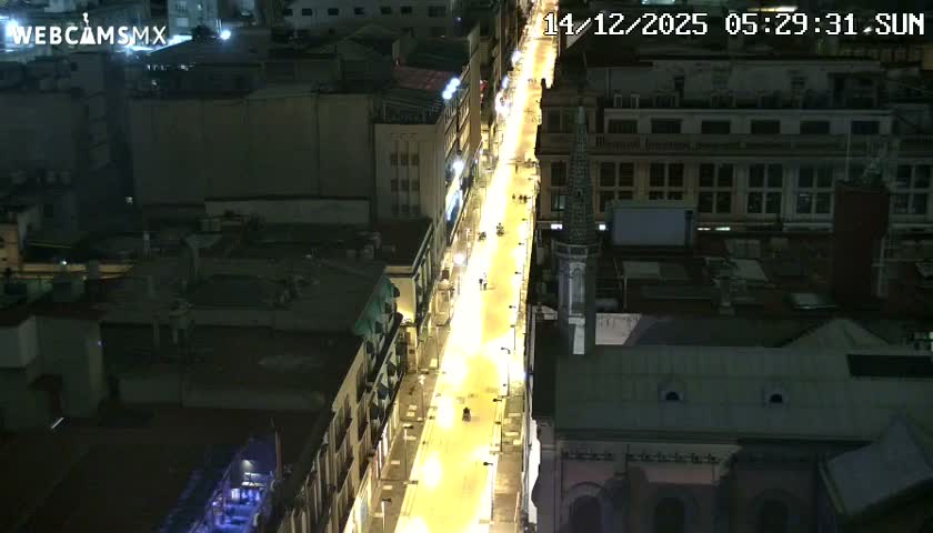A nighttime aerial view reveals a brightly lit street flanked by buildings in a city, with a spire visible, under clear skies.