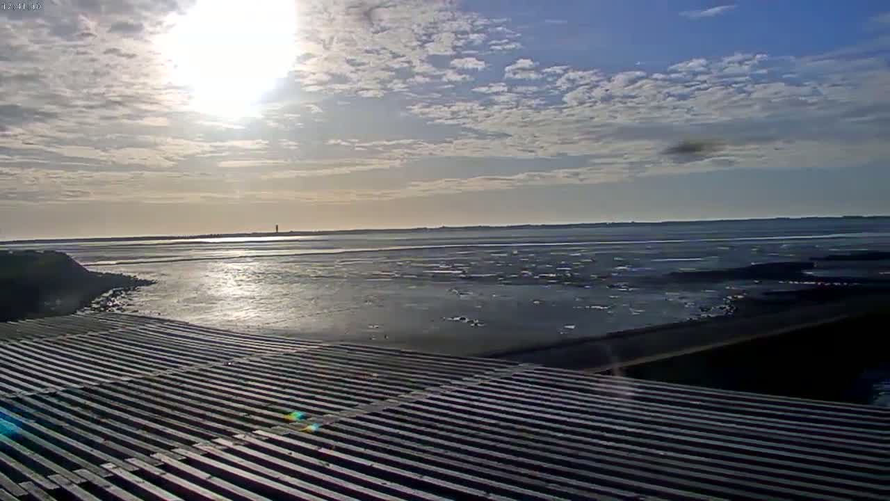 A mostly sunny day shows a calm expanse of water reflecting sunlight, a dark shoreline, and a slatted structure in the foreground.