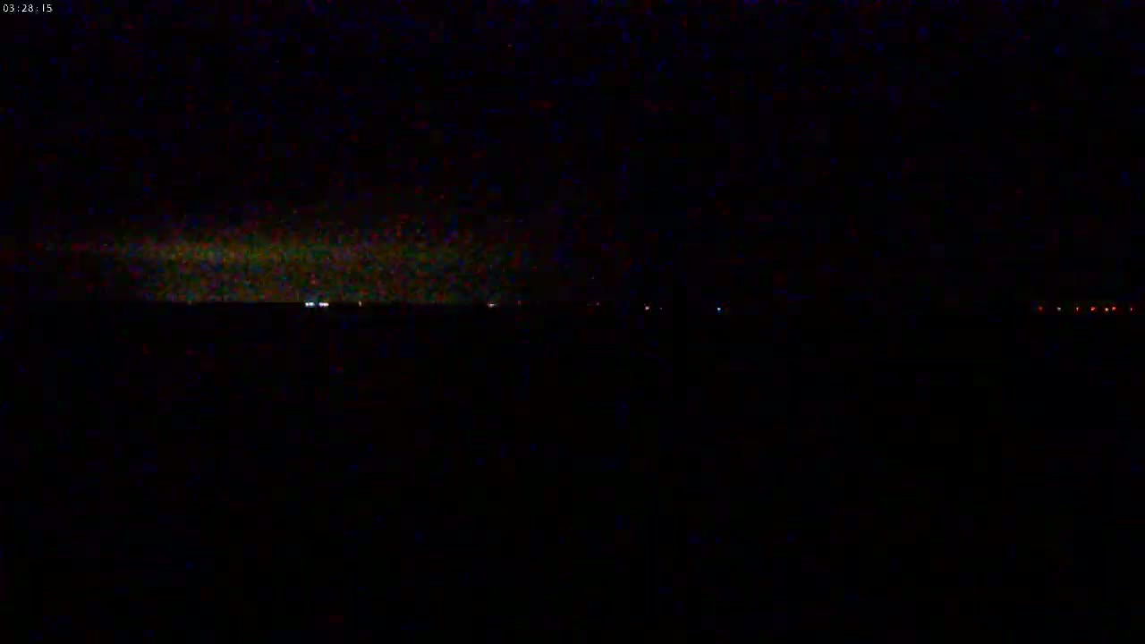A dark, nighttime landscape shows a faint, greenish glow on the horizon above a line of distant, small lights.