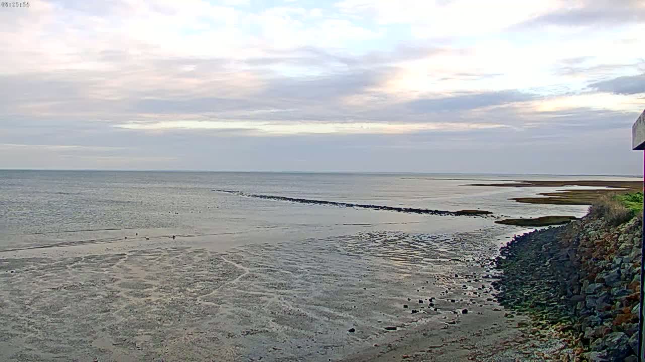 A mostly cloudy sky overlooks a calm, expansive body of water receding to reveal a muddy, textured shoreline and a rocky embankment.