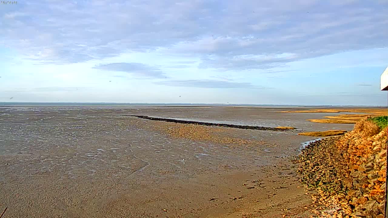 A wide expanse of mudflats stretches to a calm sea under a mostly clear, lightly cloudy sky.