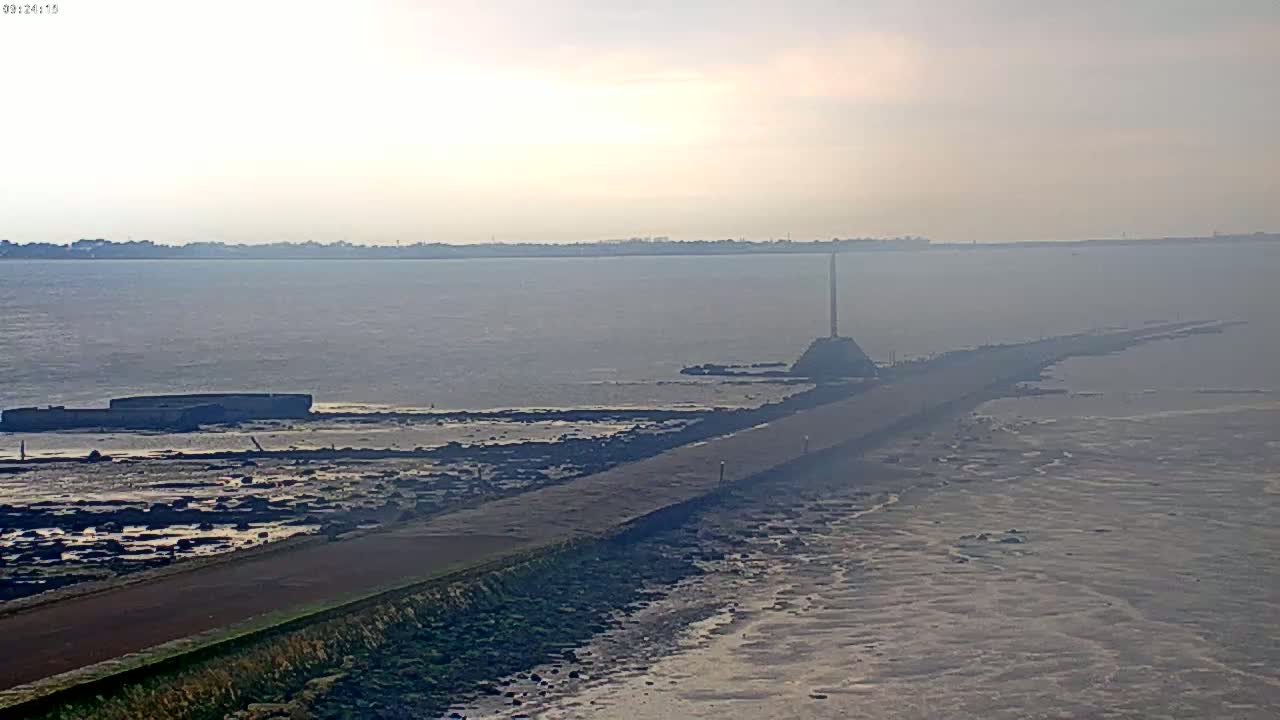 A long, narrow road extends from the foreground into a calm, hazy body of water, ending near a tall, thin structure on a small island.